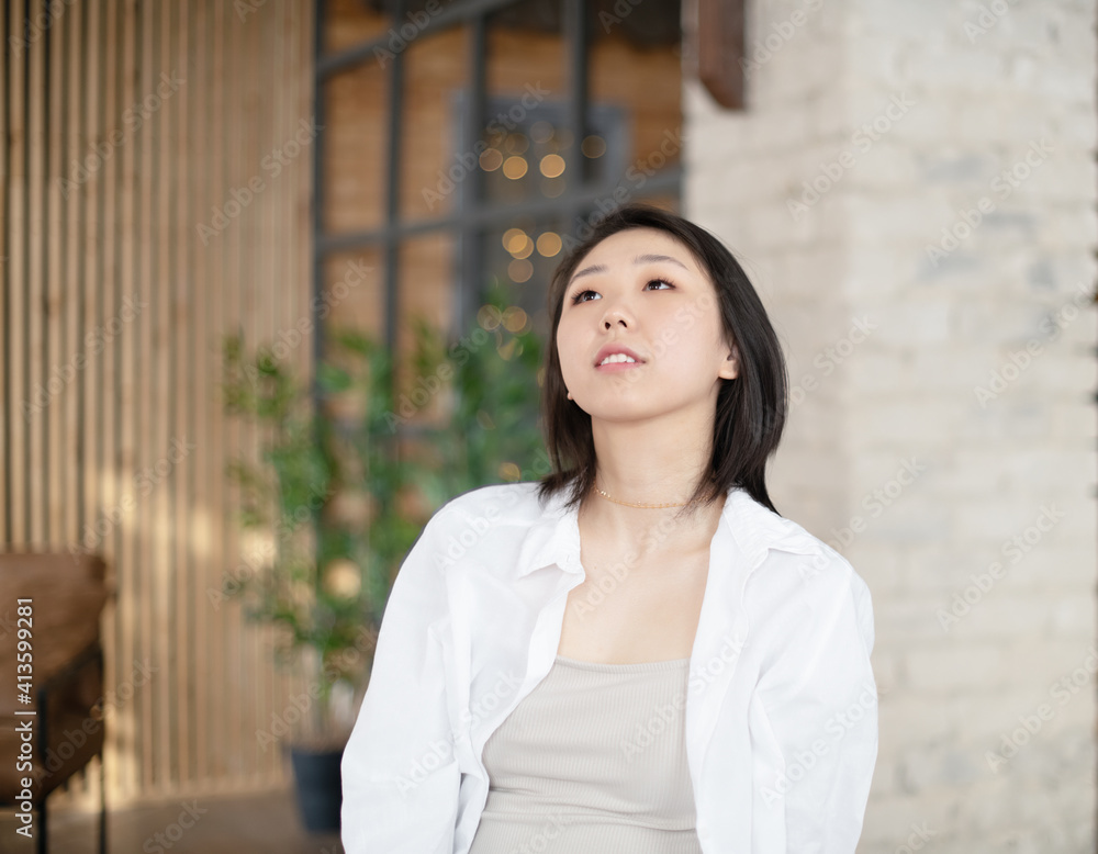 Portrait of a beautiful Asian young woman in a white fashionable shirt at home in a dark loft design interior. Portrait of a beautiful Asian young woman in a white fashionable shirt at home in a dark loft design interior.