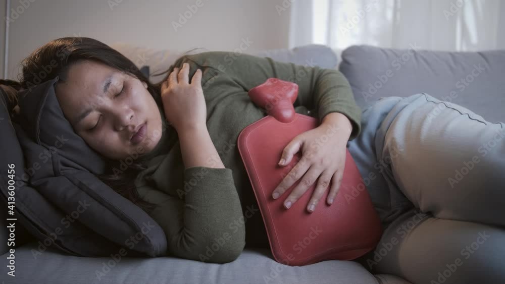 Sick woman with heavy menstrual cramps is applying a hot water bag on her stomach to relieve the pain and feel better on the living room sofa Sick woman with heavy menstrual cramps is applying a hot water bag on her stomach to relieve the pain and feel better on the living room sofa