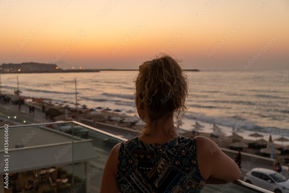 Fototapeta premium Blond girl with curly hair looking towards Old Venetian harbor and beach with umbrellas in the evening in city of Rethymno, Crete, Greece
