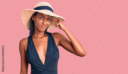 Young african american woman wearing swimsuit and summer hat pointing unhappy to pimple on forehead, ugly infection of blackhead. acne and skin problem