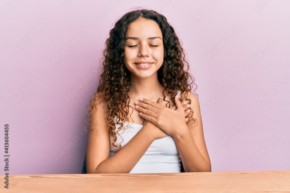 Teenager hispanic girl wearing casual clothes sitting on the table smiling with hands on chest, eyes closed with grateful gesture on face. health concept. Teenager hispanic girl wearing casual clothes sitting on the table smiling with hands on chest, eyes closed with grateful gesture on face. health concept.