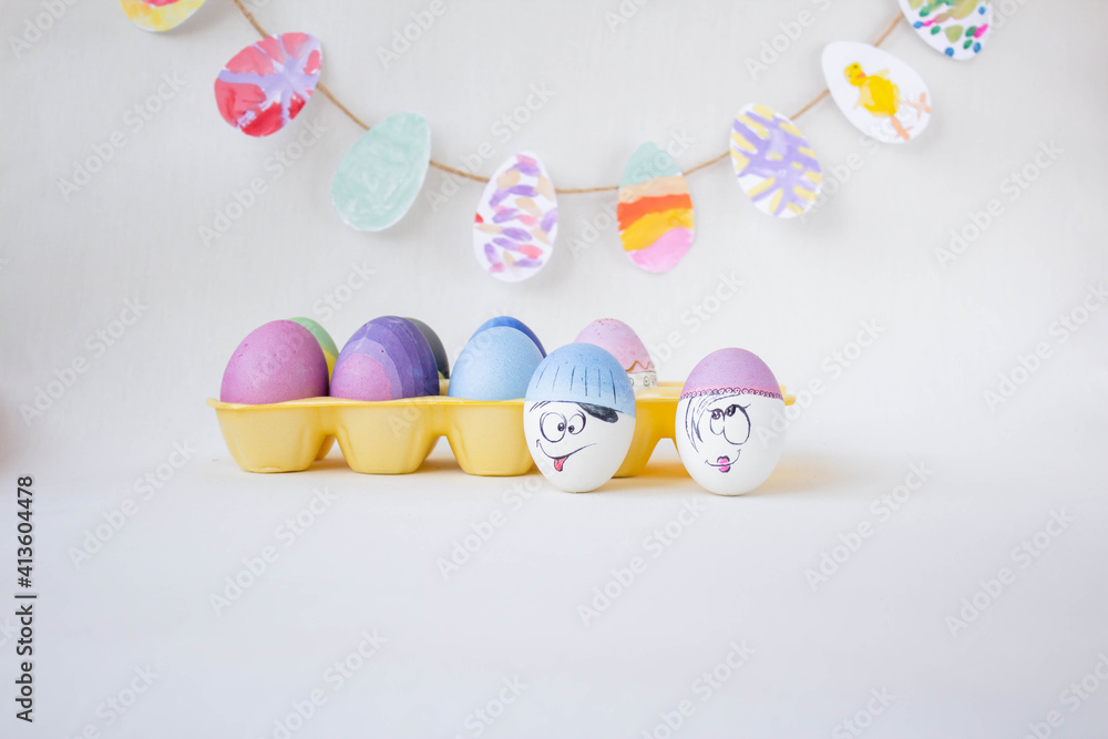 Multi-colored eggs on a yellow stand. Holiday and joy of light Easter. Hand-painted blue, blue, pink eggs. White eggs with funny faces on a white background. Easter garland.