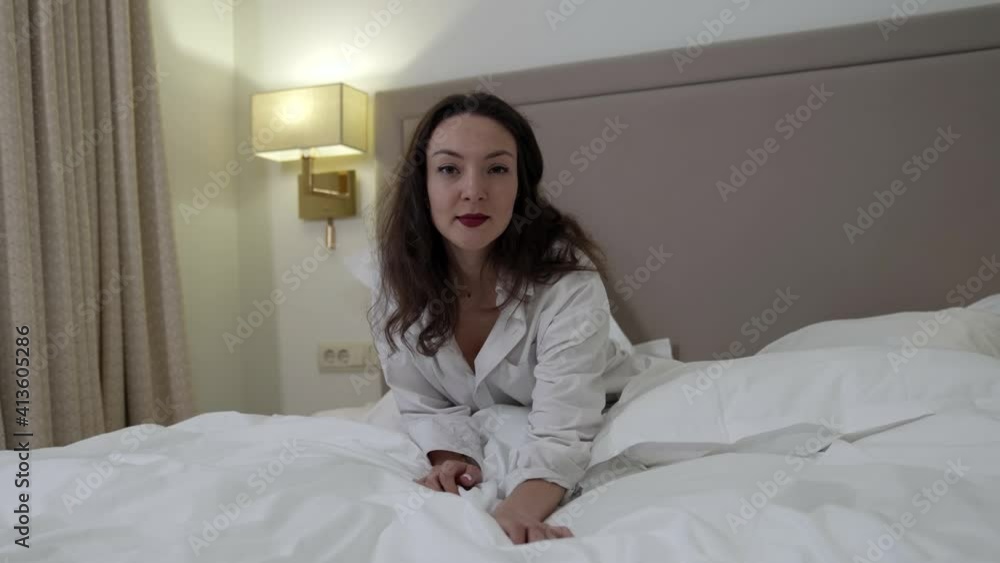 A cheerful brunette woman in a white shirt playfully poses in a room on a bed on white bed linen. A cheerful brunette woman in a white shirt playfully poses in a room on a bed on white bed linen.