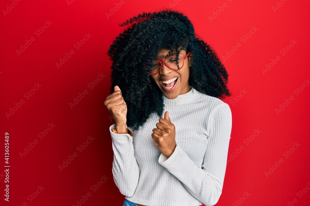 African american woman with afro hair wearing casual sweater and glasses celebrating surprised and amazed for success with arms raised and eyes closed African american woman with afro hair wearing casual sweater and glasses celebrating surprised and amazed for success with arms raised and eyes closed