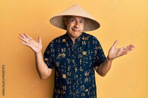 Middle age bald man wearing traditional asian straw hat clueless and confused with open arms, no idea and doubtful face.