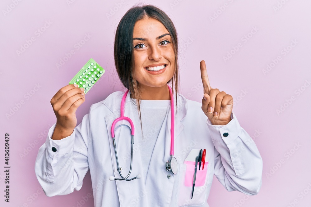 Beautiful hispanic woman wearing doctor uniform holding prescription pills smiling with an idea or question pointing finger with happy face, number one Beautiful hispanic woman wearing doctor uniform holding prescription pills smiling with an idea or question pointing finger with happy face, number one