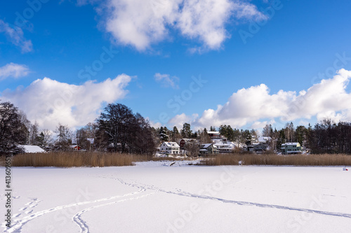 A snow covered field on the edge of village in northern Europe. Footprints of people in the snow. In the distance rural houses. Blue sky background.