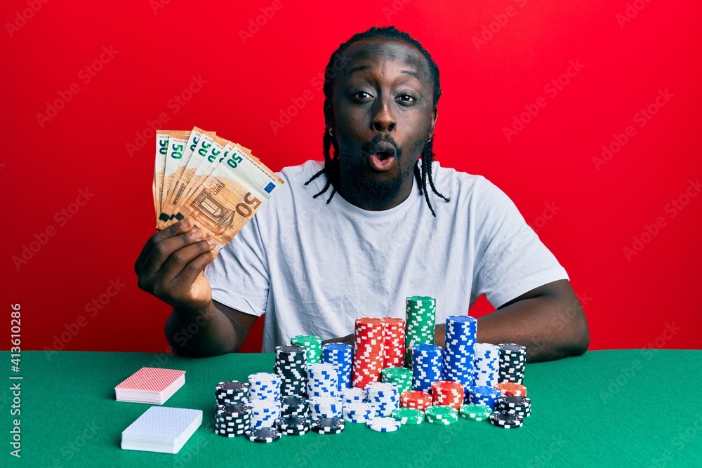 Handsome young black man playing poker holding 50 euros banknotes scared and amazed with open mouth for surprise, disbelief face Handsome young black man playing poker holding 50 euros banknotes scared and amazed with open mouth for surprise, disbelief face
