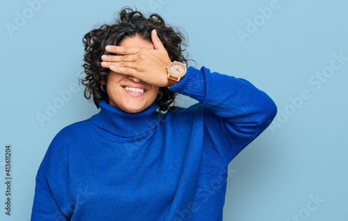 Young hispanic woman with curly hair wearing turtleneck sweater smiling and laughing with hand on face covering eyes for surprise. blind concept.