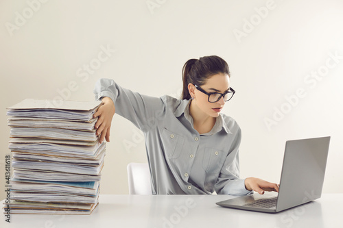 Young accountant working on laptop computer sitting at desk with pile of papers. Paperwork vs electronic documents. Storing files in digital database. Having quick convenient access to storage system photo