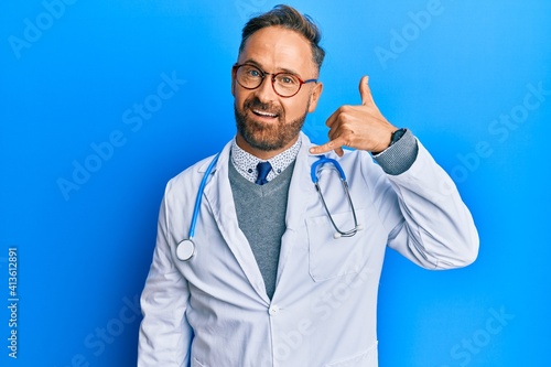 Handsome middle age man wearing doctor uniform and stethoscope smiling doing phone gesture with hand and fingers like talking on the telephone. communicating concepts.