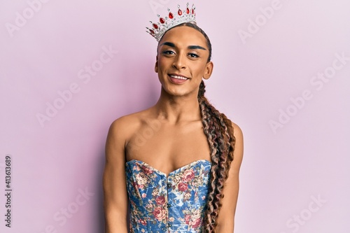 Hispanic man wearing make up and long hair wearing princess crown looking positive and happy standing and smiling with a confident smile showing teeth