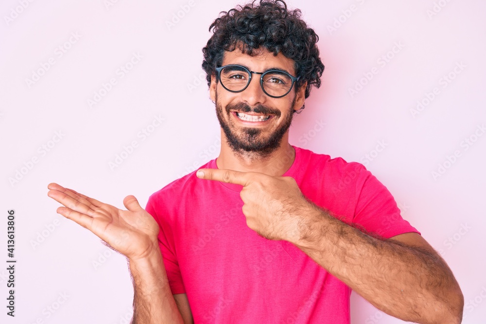 Handsome young man with curly hair and bear wearing casual clothes and glasses amazed and smiling to the camera while presenting with hand and pointing with finger. Handsome young man with curly hair and bear wearing casual clothes and glasses amazed and smiling to the camera while presenting with hand and pointing with finger.