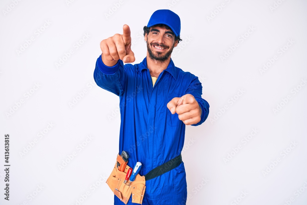 Handsome young man with curly hair and bear weaing handyman uniform pointing to you and the camera with fingers, smiling positive and cheerful
