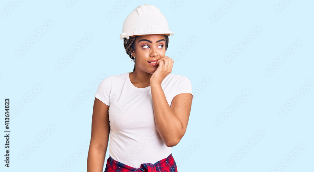 Young african american woman with braids wearing hardhat and builder clothes looking stressed and nervous with hands on mouth biting nails. anxiety problem. Young african american woman with braids wearing hardhat and builder clothes looking stressed and nervous with hands on mouth biting nails. anxiety problem.