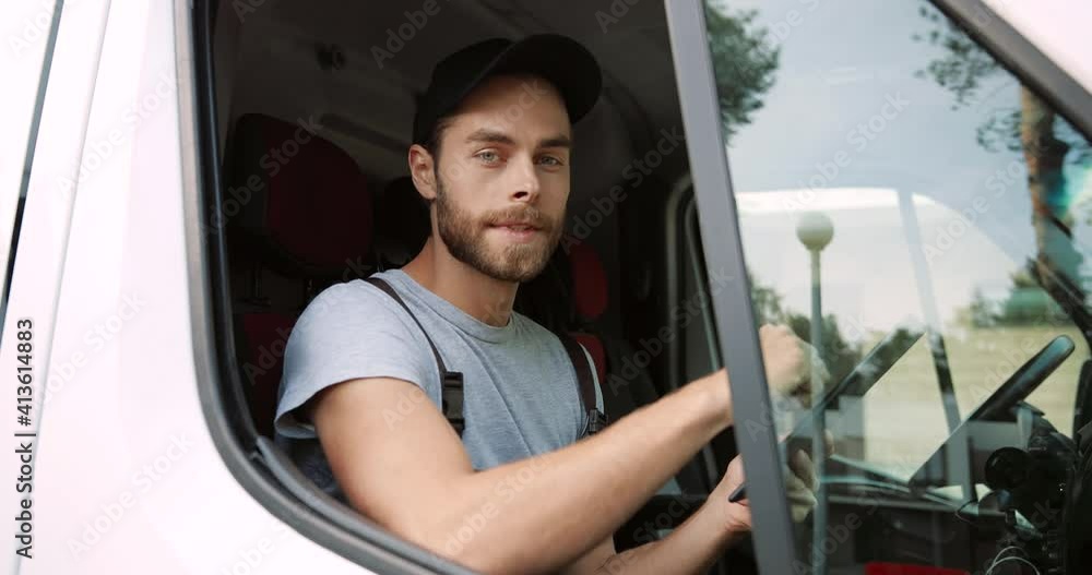 Side view. Close up of Caucasian young joyful handsome male courier sitting on front passenger seat in van tapping and browsing online on tablet device smiling to camera. Delivering orders. Side view. Close up of Caucasian young joyful handsome male courier sitting on front passenger seat in van tapping and browsing online on tablet device smiling to camera. Delivering orders.