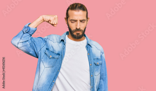 Attractive man with long hair and beard wearing casual denim jacket strong person showing arm muscle, confident and proud of power