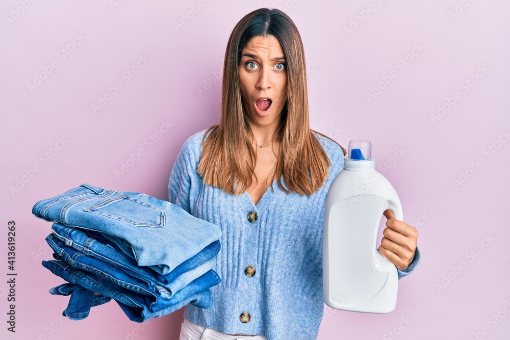 Brunette young woman holding jeans for laundry and detergent bottle afraid and shocked with surprise and amazed expression, fear and excited face. Brunette young woman holding jeans for laundry and detergent bottle afraid and shocked with surprise and amazed expression, fear and excited face.
