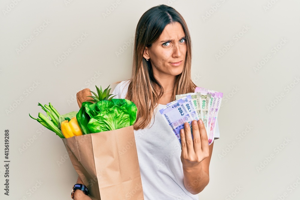 Brunette young woman holding groceries and indian rupee banknotes in shock face, looking skeptical and sarcastic, surprised with open mouth Brunette young woman holding groceries and indian rupee banknotes in shock face, looking skeptical and sarcastic, surprised with open mouth
