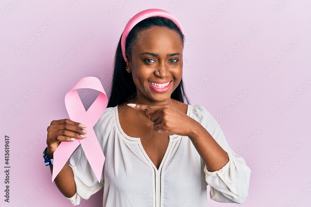 Young african woman holding pink cancer ribbon smiling happy pointing with hand and finger Young african woman holding pink cancer ribbon smiling happy pointing with hand and finger