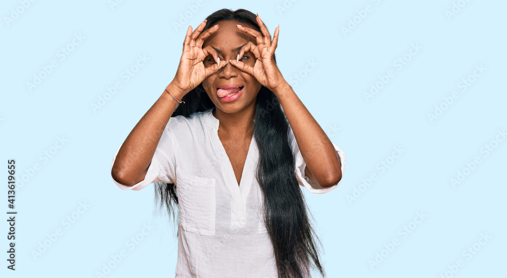 Young black woman wearing casual clothes doing ok gesture like binoculars sticking tongue out, eyes looking through fingers. crazy expression. Young black woman wearing casual clothes doing ok gesture like binoculars sticking tongue out, eyes looking through fingers. crazy expression.