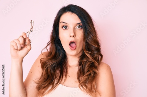 Young beautiful brunette woman holding eyelash curler scared and amazed with open mouth for surprise, disbelief face