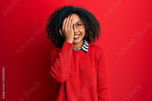 Beautiful african american woman with afro hair wearing sweater and glasses covering one eye with hand, confident smile on face and surprise emotion.