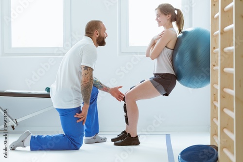 Handsome tattooed trainer assisting a girl during physiotherapy exercises with ball