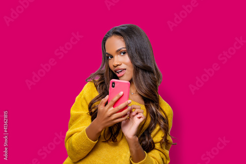 Hello. Portrait of cool cheerful African American girl in yellow sweater having video-call or taking selfie, holding smart phone in hand shooting selfie on front camera isolated on pink background.   