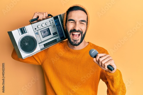 Young hispanic man holding boombox, listening to music singing with microphone smiling and laughing hard out loud because funny crazy joke.