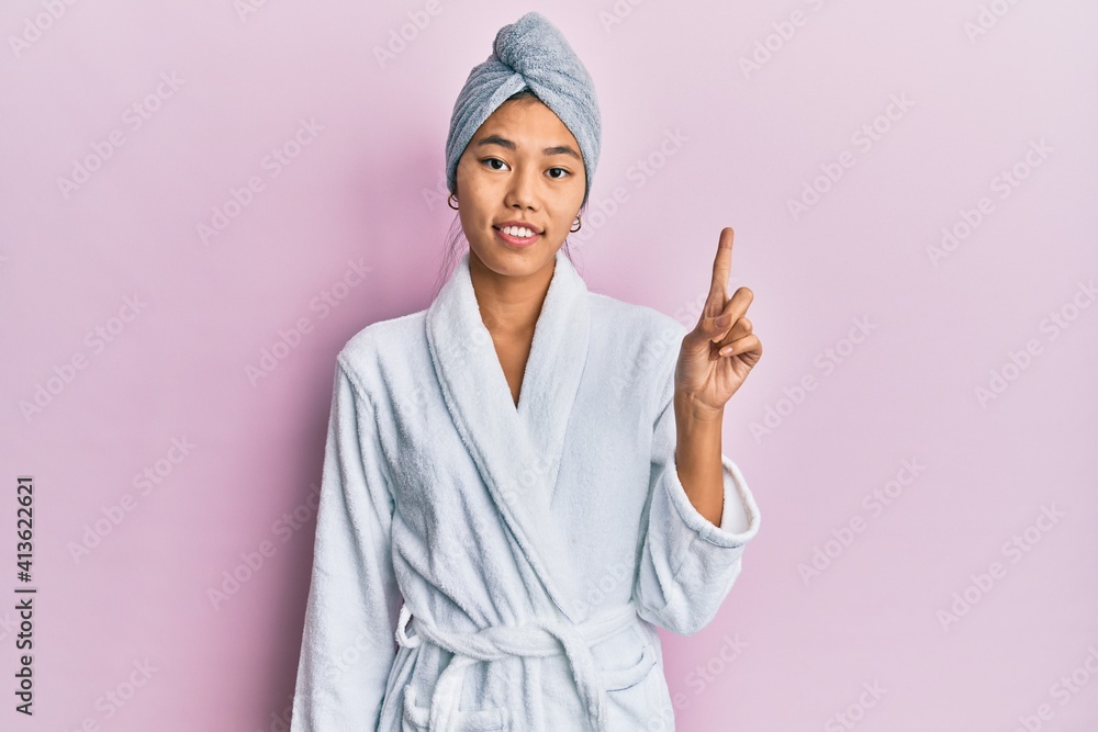 Young chinese woman wearing shower towel cap and bathrobe showing and pointing up with finger number one while smiling confident and happy. Young chinese woman wearing shower towel cap and bathrobe showing and pointing up with finger number one while smiling confident and happy.