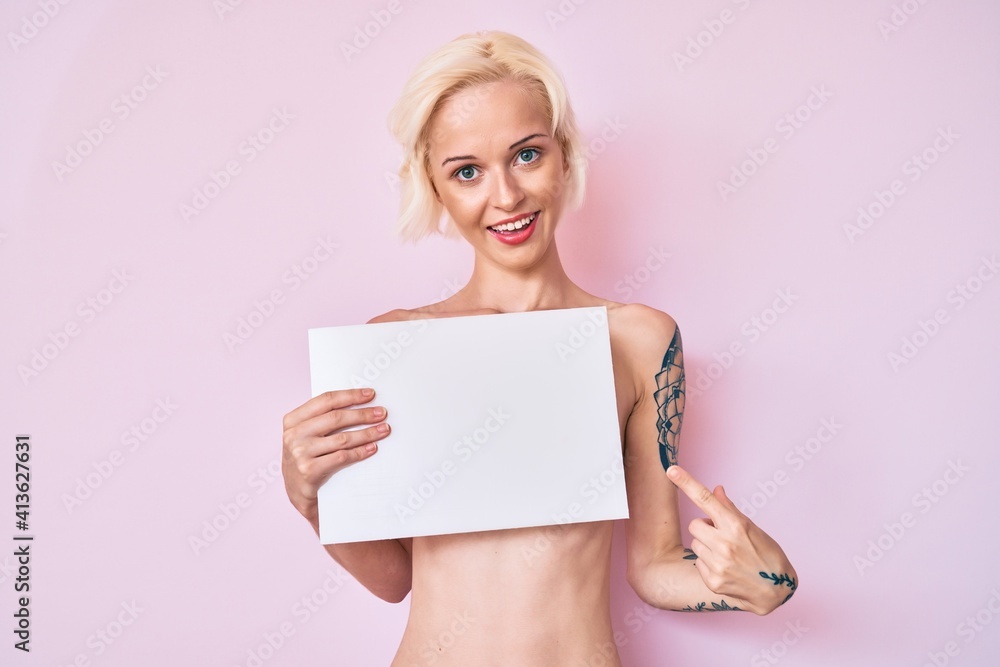 Young blonde woman with tattoo standing shirtless holding empty blank banner smiling happy pointing with hand and finger Young blonde woman with tattoo standing shirtless holding empty blank banner smiling happy pointing with hand and finger