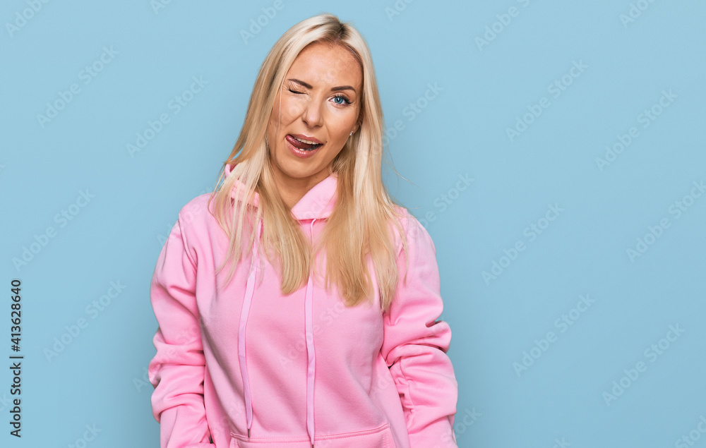 Young blonde woman wearing casual sweatshirt winking looking at the camera with sexy expression, cheerful and happy face. Young blonde woman wearing casual sweatshirt winking looking at the camera with sexy expression, cheerful and happy face.