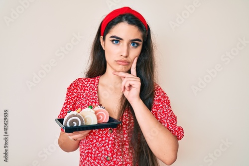 Brunette teenager girl holding cake sweets serious face thinking about question with hand on chin, thoughtful about confusing idea
