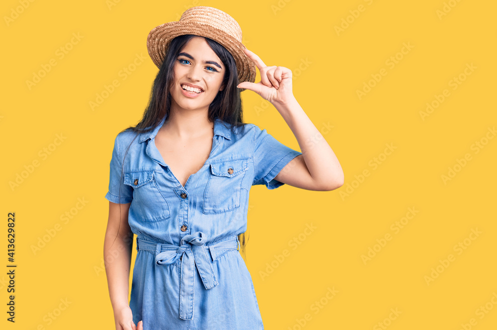 Brunette teenager girl wearing summer hat smiling and confident gesturing with hand doing small size sign with fingers looking and the camera. measure concept. Brunette teenager girl wearing summer hat smiling and confident gesturing with hand doing small size sign with fingers looking and the camera. measure concept.