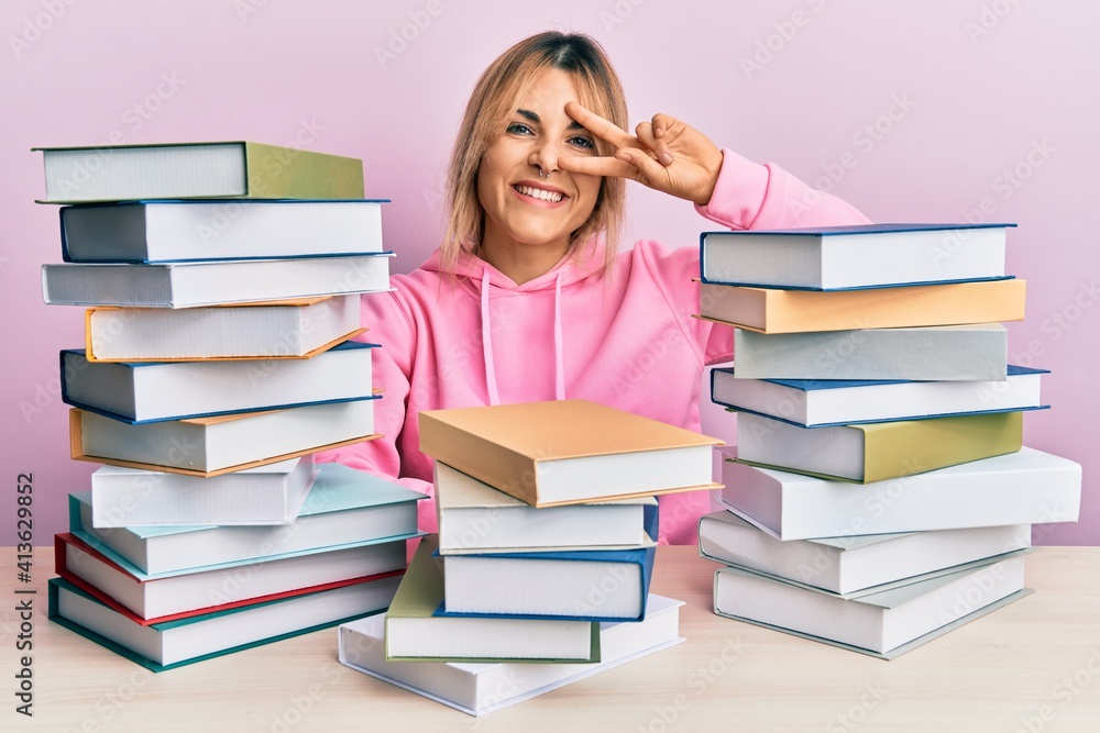 Young caucasian woman sitting on the table with books doing peace symbol with fingers over face, smiling cheerful showing victory Young caucasian woman sitting on the table with books doing peace symbol with fingers over face, smiling cheerful showing victory