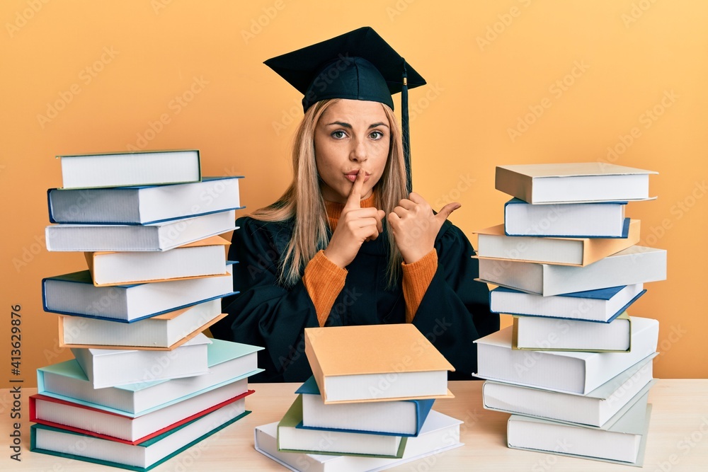 Young caucasian woman wearing graduation ceremony robe sitting on the table asking to be quiet with finger on lips pointing with hand to the side. silence and secret concept.