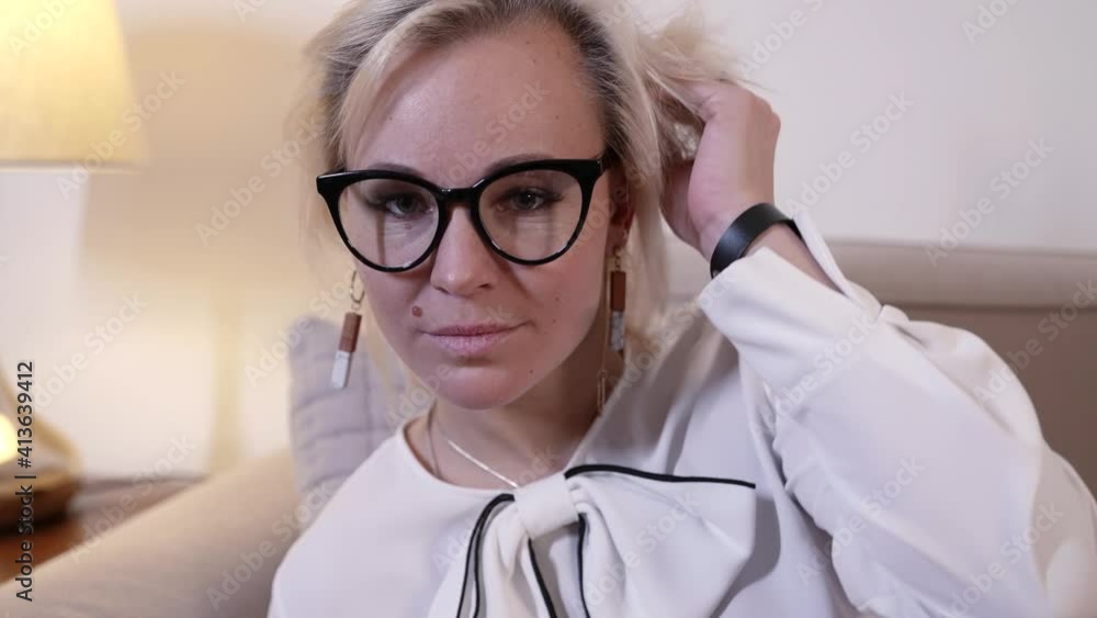 Portrait of a blonde woman with glasses is in the room, on the sofa and posing, wearing a white shirt. Portrait of a blonde woman with glasses is in the room, on the sofa and posing, wearing a white shirt.