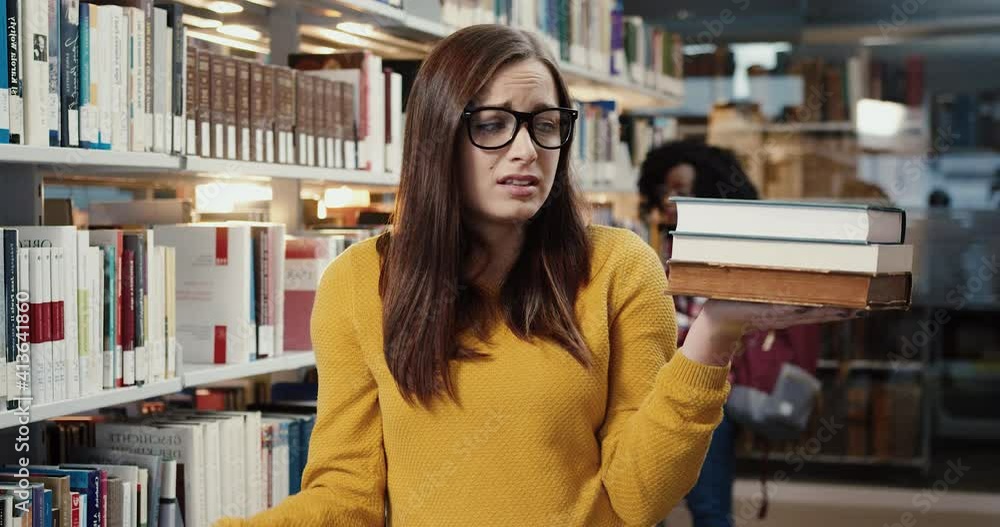 Portrait of unfortunate student stands in library and fearfully examines the number of books that need to be read quickly. Portrait of unfortunate student stands in library and fearfully examines the number of books that need to be read quickly.