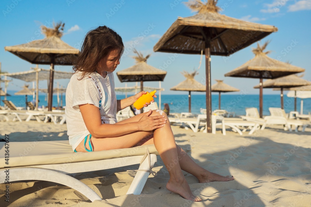 Mature woman sitting onsun lounger on sandy beach applying sunscreen to her leg
