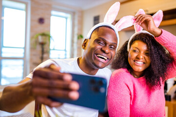 Young african american man and woman wearing cute easter bunny ears in the house taking selfie on smartphone
