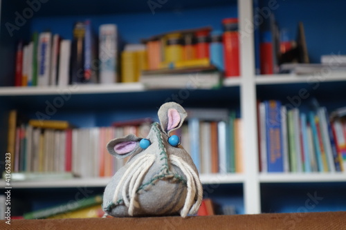 Slebstgebastelte Stoffspielzeug Maus vor buntem chaotischen Bücherregal. Lesemaus mit blauen Augen, Schnurrbarthaaren und treuem Hundeblick.