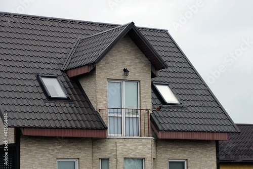 open iron balcony on the gray brick wall of the house under the brown tiled roof against the sky
