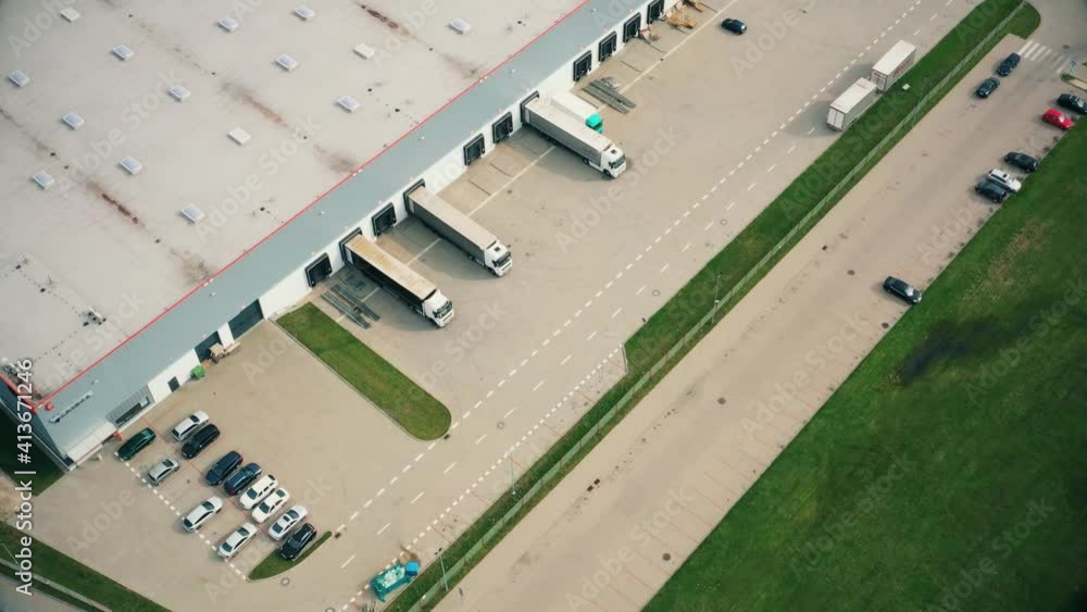 Aerial following shot of semitrailer trucks travelling along a warehouse on the parking lot in