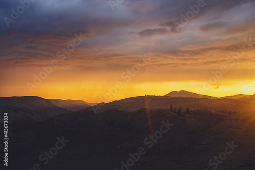 Atmospheric landscape with silhouettes of mountains with trees on background of dawn sky with vivid orange sunlight and sun rays. Colorful nature scenery with sunset or sunrise of illuminating color.