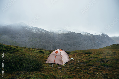 Scenic alpine landscape with tent on green hill among rocks. Beautiful snow-capped mountains in low clouds. Atmospheric scenery with tent in highlands in overcast weather. High mountains covered snow.