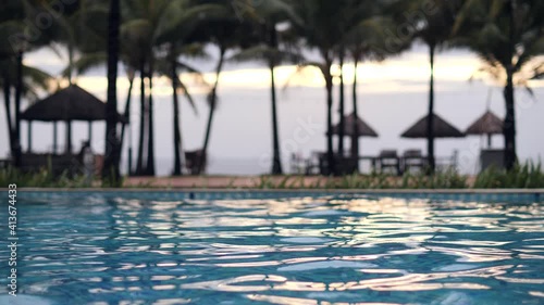 A view of a swimming pool in resort with unfocused coconut trees and beach huts