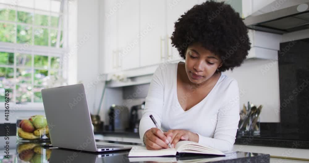 African american woman taking notes and using laptop in the kitchen while working from home African american woman taking notes and using laptop in the kitchen while working from home