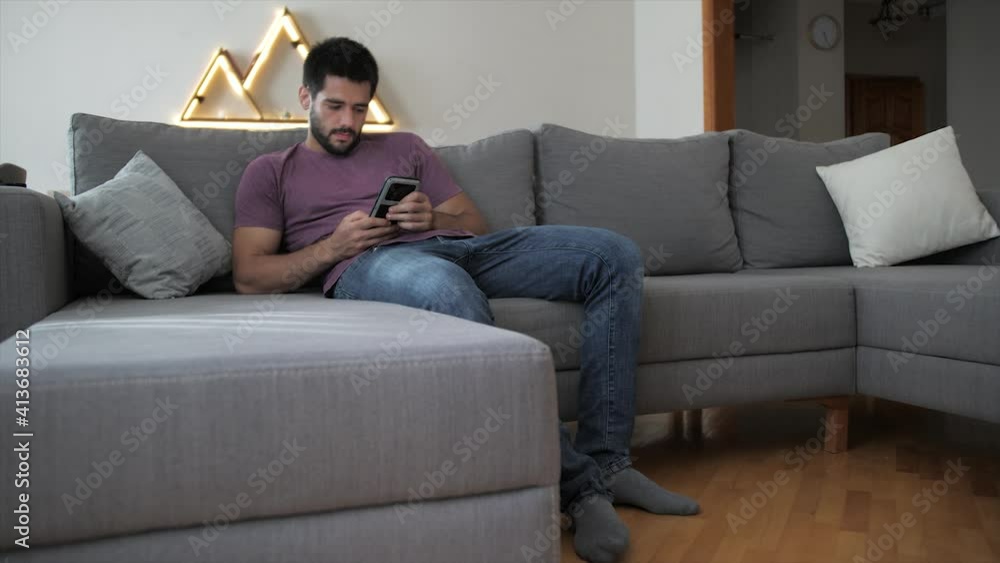 Relaxed young man in purple t-shirt and blue jeans sitting on the grey couch in a cozy living room alone and surfing social media on mobile phone Relaxed young man in purple t-shirt and blue jeans sitting on the grey couch in a cozy living room alone and surfing social media on mobile phone
