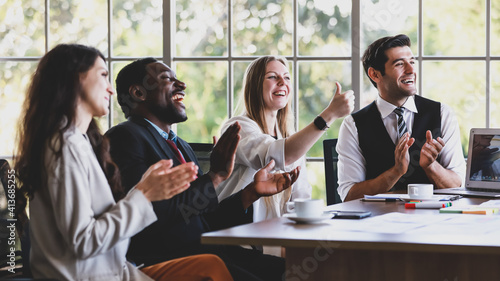 Group of four professional multiethnic businessmen and businesswomen sitting, clapping hands and showing thump up gesture to celebrate the new project in a company meeting at office working table